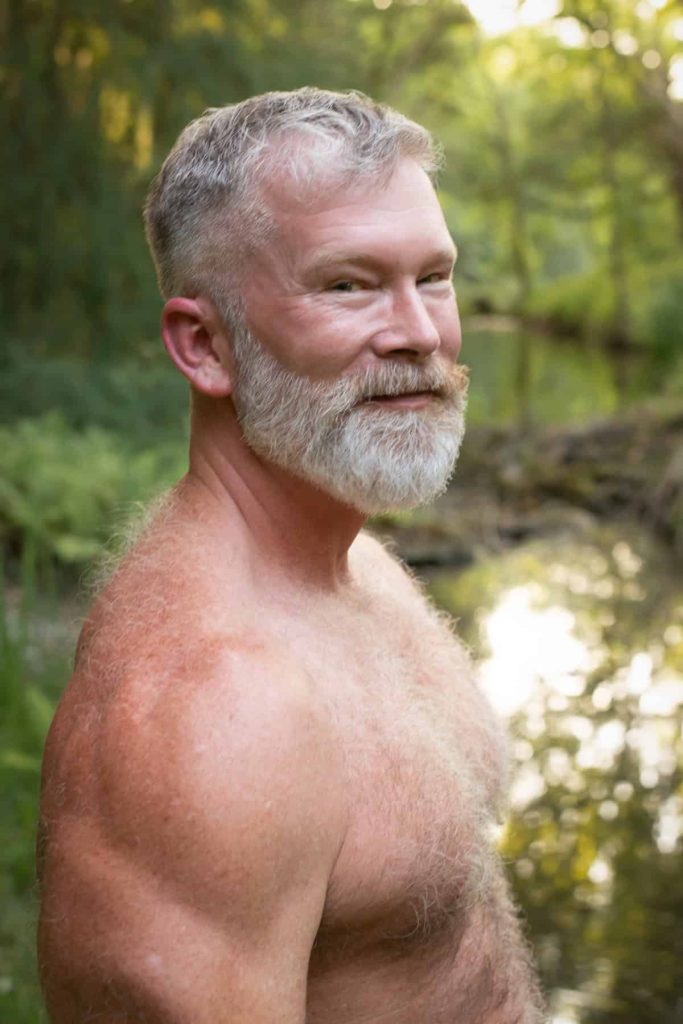 Shirtless, furry man outdoors by a stream.